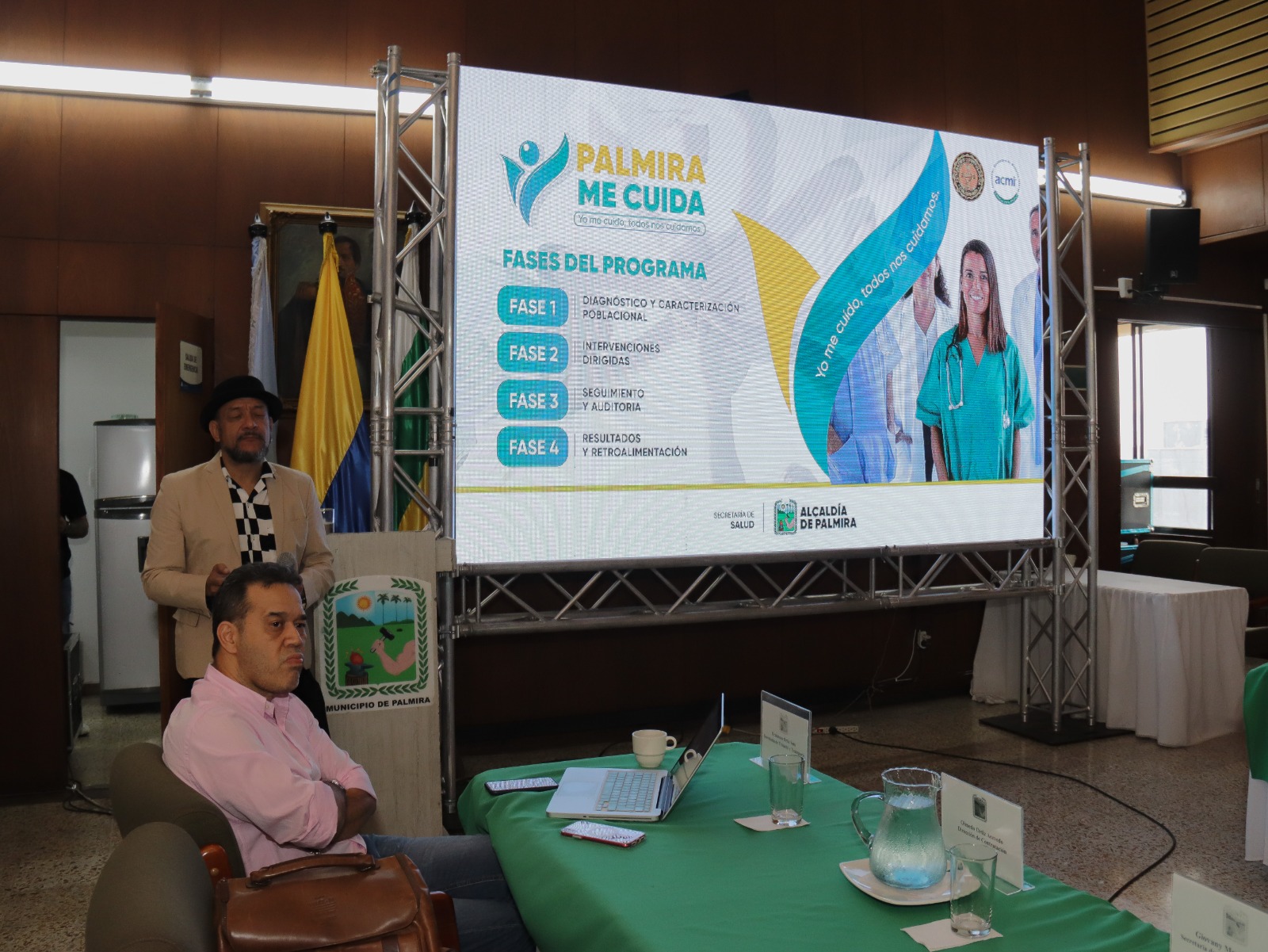 Presentación del programa institucional 'Palmira Me Cuida'. Una pantalla grande muestra las fases del proyecto junto a imágenes de personal médico. En primer plano, personas asistentes escuchan la exposición en un salón institucional