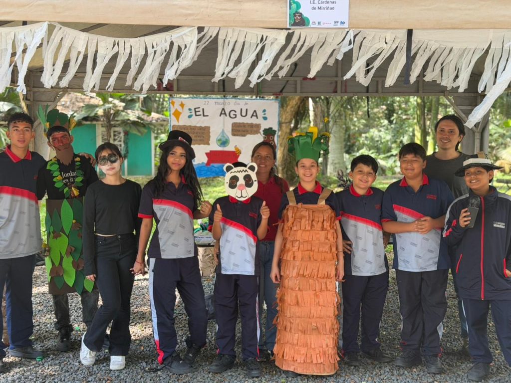 Un grupo de niños y niñas con disfraces creativos hechos a mano que representan la naturaleza (árboles, hojas, un panda, un tronco) posan sonriendo junto a sus profesores frente a una carpa con un cartel que dice "22 marzo día del EL AGUA".