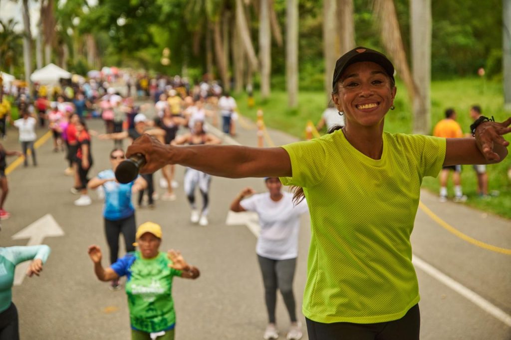 Una mujer sonriente en primer plano con una camiseta verde neón lidera una clase de ejercicio al aire libre en una calle llena de gente