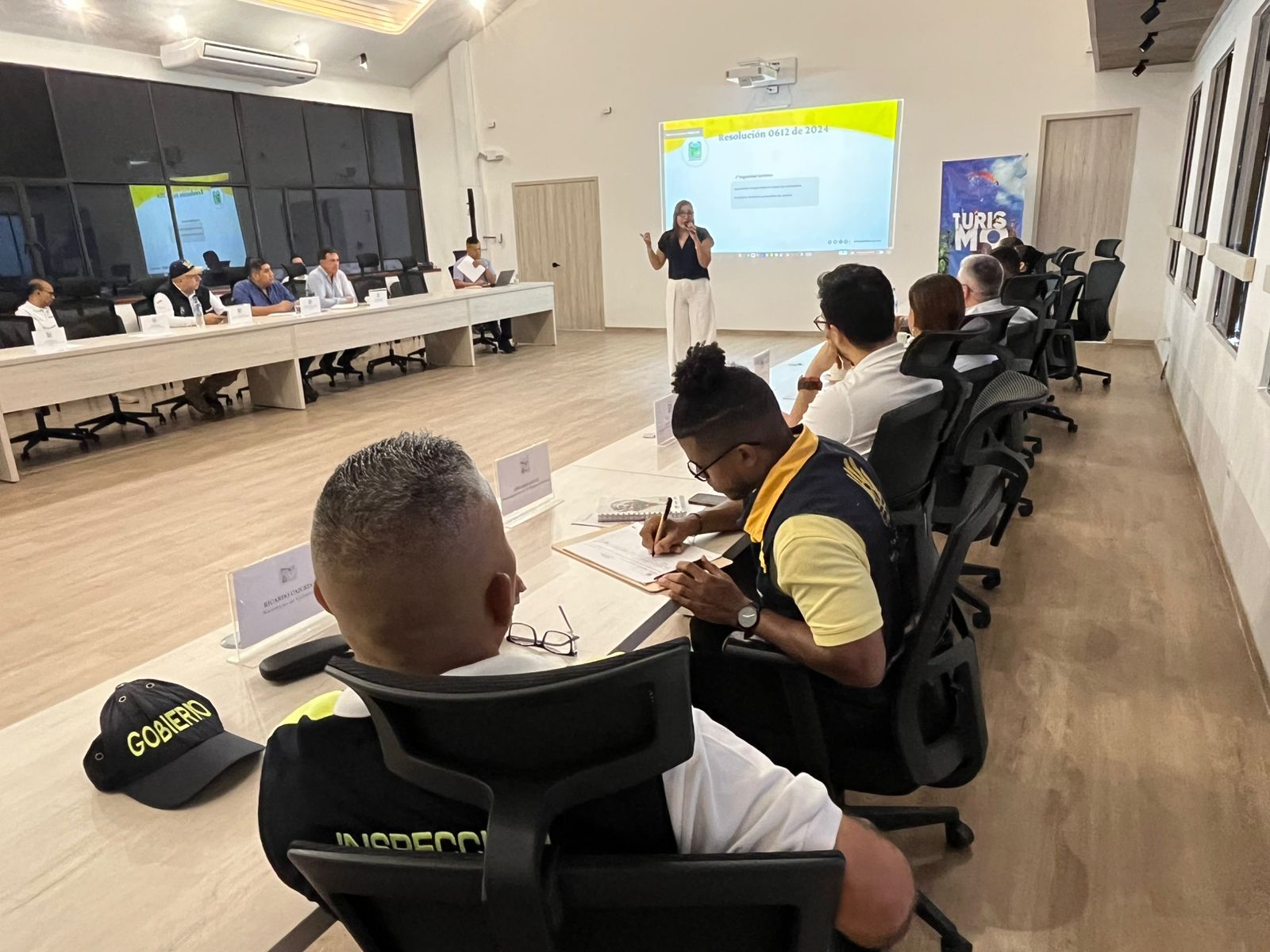 Grupo de 12 profesionales y ciudadanos sentados alrededor de una gran mesa de madera, analizando proyectos educativos frente a una pantalla con logotipos institucionales en una moderna sala de la Alcaldía de Palmira.