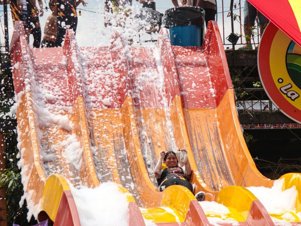Una joven sonríe mientras se desliza por un gran tobogán inflable de colores naranja y rojo cubierto de espuma blanca durante un festival al aire libre.
