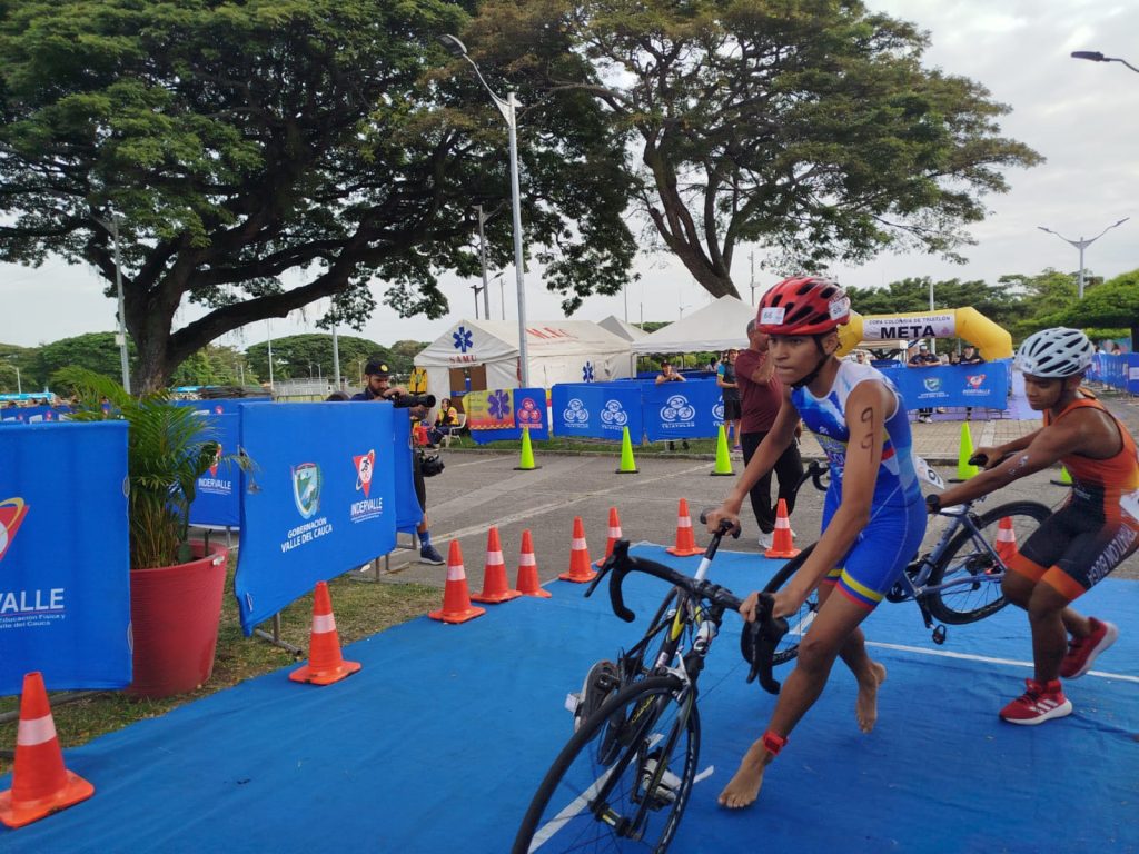 Dos jóvenes triatletas corren en paralelo sobre una alfombra azul en una calle de Palmira, con un competidor de primer plano empujando su bicicleta de ruta mientras el otro corre a su lado.