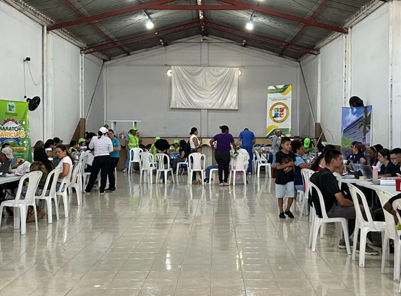 Amplio salón blanco con filas de mesas donde ciudadanos reciben atención de salud y servicios administrativos por parte de funcionarios de la Alcaldía de Palmira.