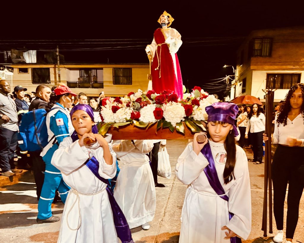 Dos niños vestidos de blanco con túnicas y capuchas moradas llevan en hombros un paso procesional de madera con la estatua de un santo en una procesión nocturna por las calles de Palmira.