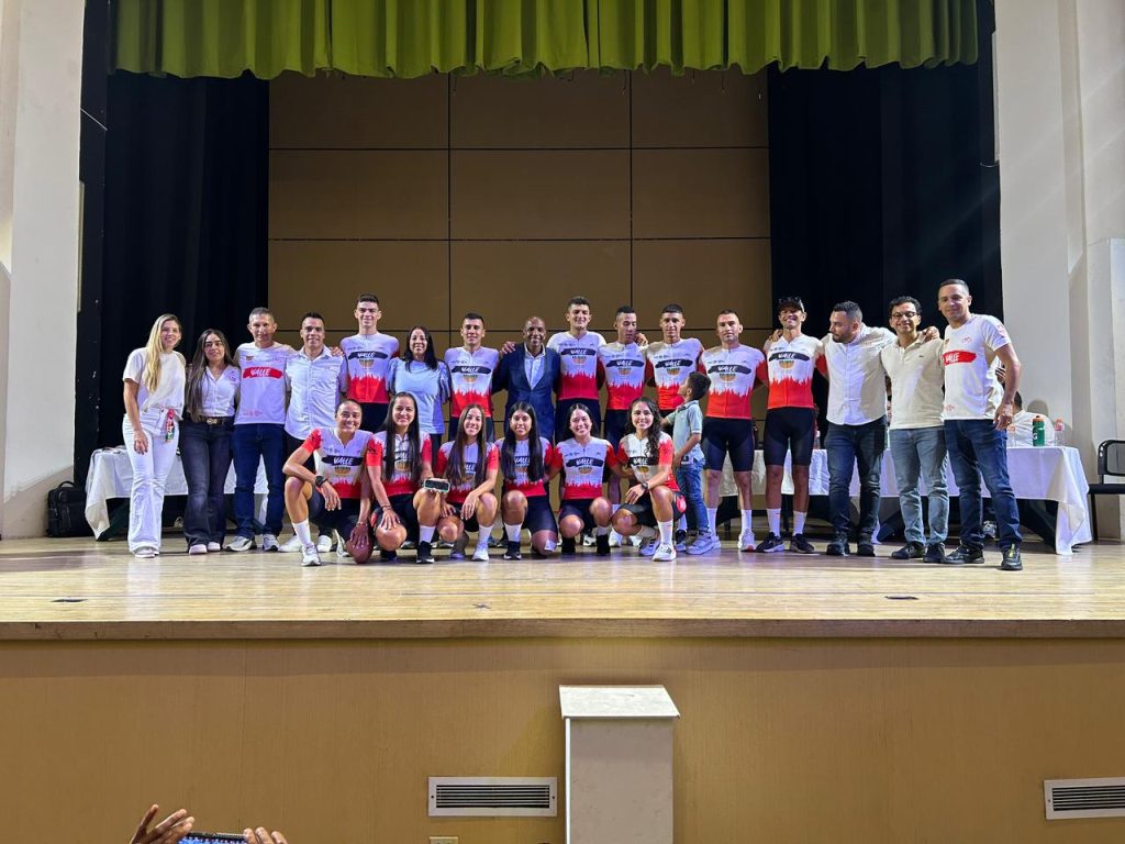 El director del IMDER Palmira, un hombre negro con gorra y camiseta negra, hace la señal de la victoria en primer plano mientras cientos de participantes vestidos de amarillo con gorras del IMDER celebran detrás en un parque arbolado.