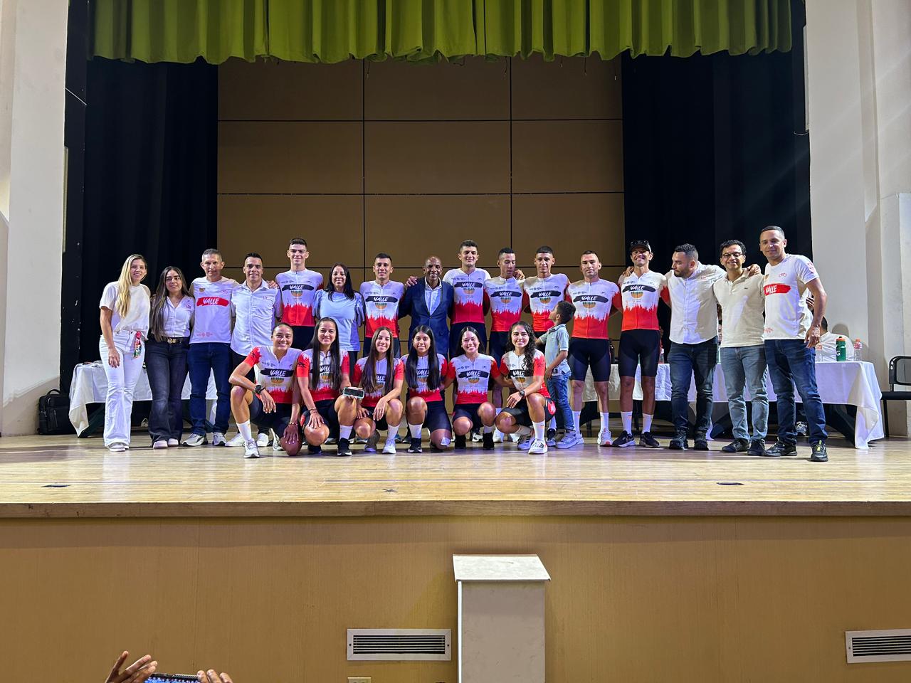 El director del IMDER Palmira, un hombre negro con gorra y camiseta negra, hace la señal de la victoria en primer plano mientras cientos de participantes vestidos de amarillo con gorras del IMDER celebran detrás en un parque arbolado.