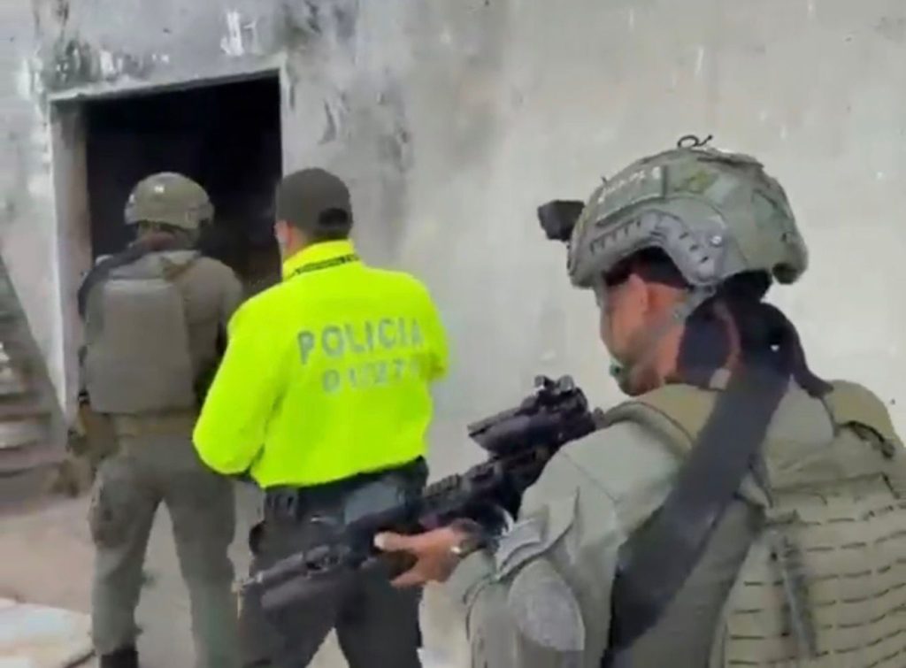 Tres oficiales de policía con uniforme táctico y equipo de protección, incluyendo cascos y chalecos, ingresan a una estructura de concreto oscuro con un oficial de uniforme amarillo brillante que dice "POLICÍA", custodiando la entrada.
