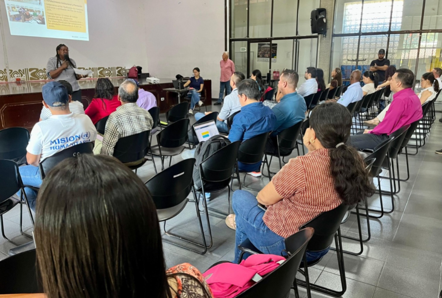 Una funcionaria de piel negra con trenzas y una camisa a rayas se dirige a una audiencia sentada en sillas negras en un auditorio, mientras un proyector muestra una presentación en el fondo.