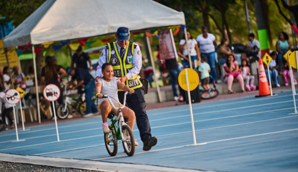 Un oficial de tránsito con uniforme amarillo y chaleco, guía a una niña con una camiseta blanca que sonríe mientras monta una bicicleta pequeña sobre una pista azul delimitada por conos naranjas y señales de tráfico circulares de "Ceda el Paso".