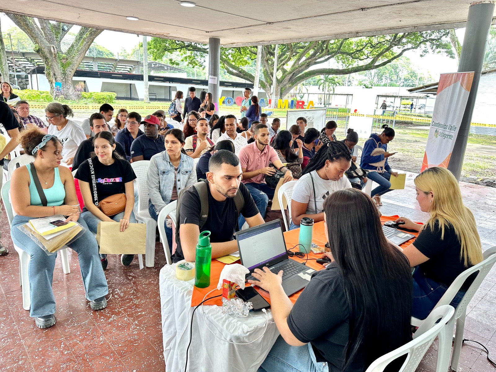 Una fila de ciudadanos espera frente a mesas de atención donde funcionarias con computadoras registran información durante una feria de empleo al aire libre.