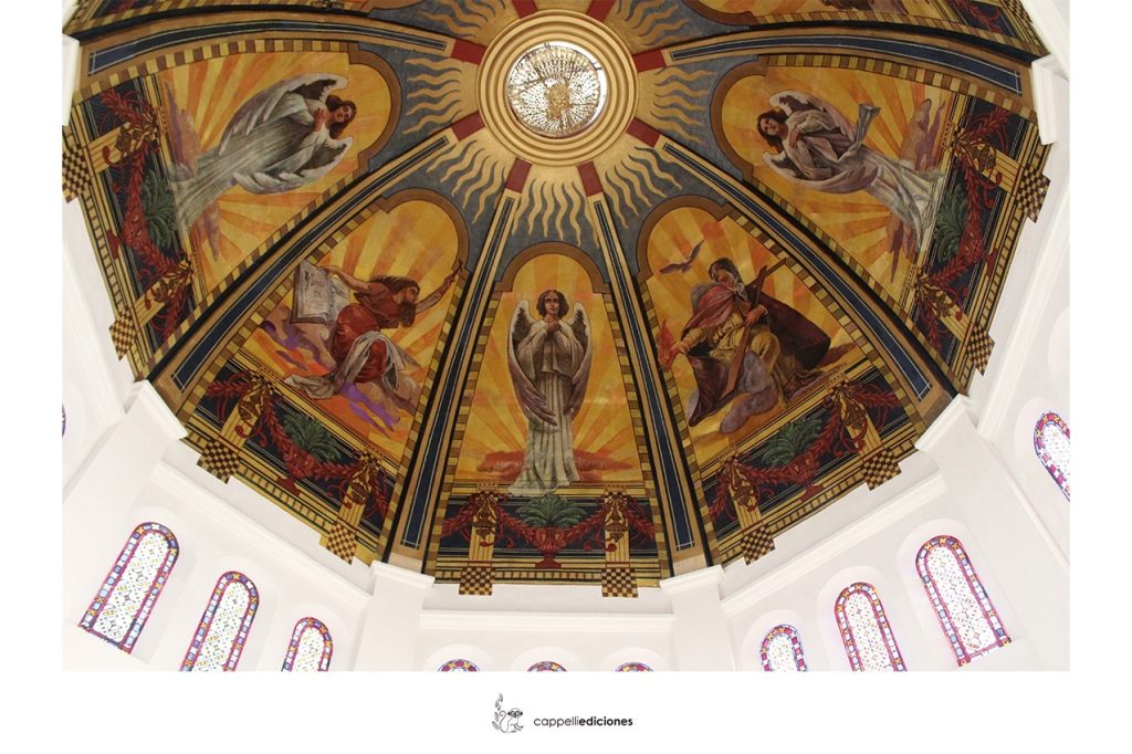 ista contrapicada de una cúpula pintada con murales de ángeles y figuras bíblicas en tonos dorados y amarillos, rodeada de vitrales coloridos.