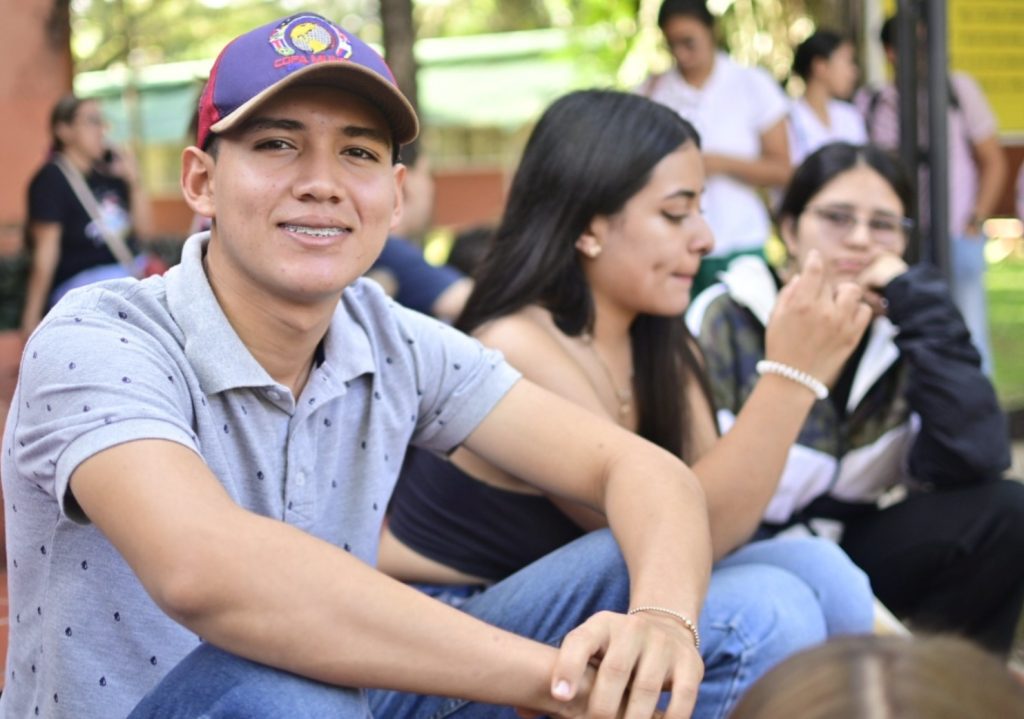 Un joven con gorra morada y camisa polo gris sonríe a la cámara mientras está sentado en el césped con otros estudiantes durante una actividad al aire libre.