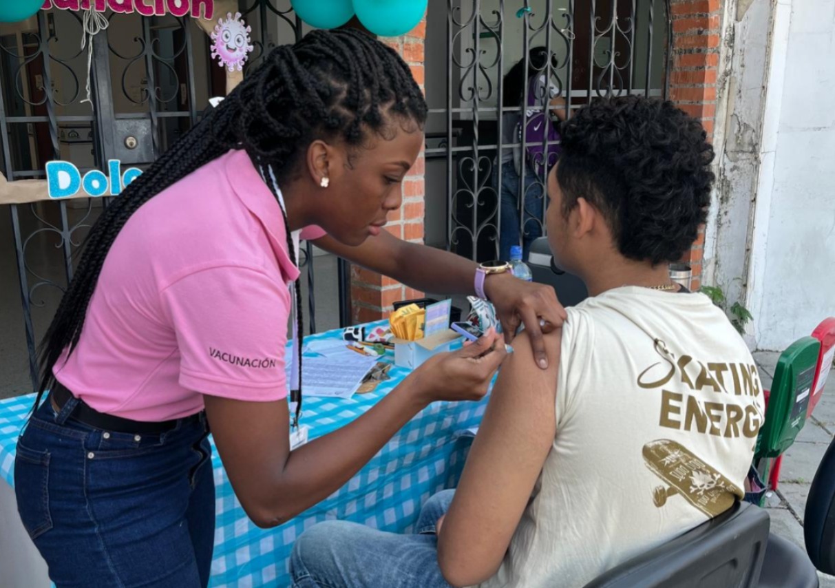 Una enfermera con una camiseta rosa que dice "VACUNACIÓN" y trenzas, administra una inyección en el hombro de un joven sentado en una silla, mientras él está de espaldas a la cámara y lleva una camiseta blanca que dice "Skating Energy".
