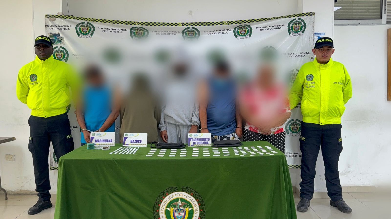 Dos integrantes de la Policía Nacional de Colombia, con chaquetas verde neón, custodian a cinco personas cuyos rostros están difuminados. Se encuentran frente a un telón institucional de la policía. Sobre una mesa con mantel verde se exhiben elementos incautados, identificados con carteles que indican "MARIHUANA", "BAZUCO" y "CLORHIDRATO DE COCAÍNA".