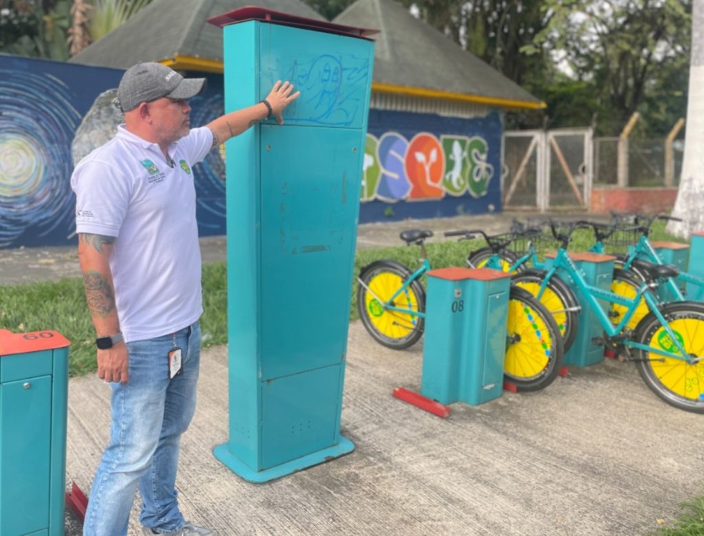 Una persona del equipo técnico de la Alcaldía de Palmira inspecciona un tótem de control de color turquesa en una estación del sistema "Palmibici". La persona viste una camiseta blanca con logotipos institucionales, gorra gris y jeans. A su derecha se encuentran varias bicicletas públicas de color turquesa con rines amarillos, estacionadas en sus respectivos anclajes. Al fondo, se observa un muro con murales coloridos y vegetación de un parque.