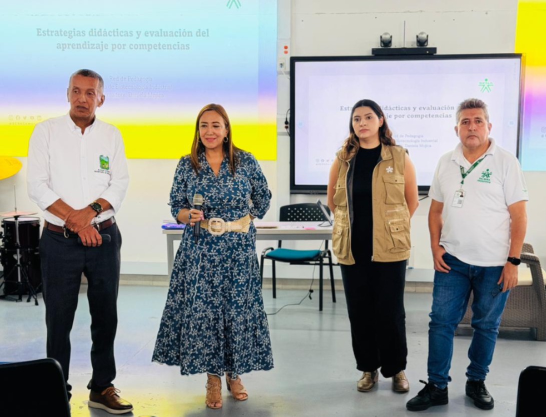 Cuatro personas, dos hombres y dos mujeres, están de pie frente a una audiencia en un salón institucional. En el fondo, dos pantallas proyectan una presentación titulada "Estrategias didácticas y evaluación del aprendizaje por competencias". La mujer en el centro-izquierda sostiene un micrófono y se dirige al público. Las personas visten uniformes institucionales de la Alcaldía de Palmira y el SENA. El ambiente es formal y educativo.