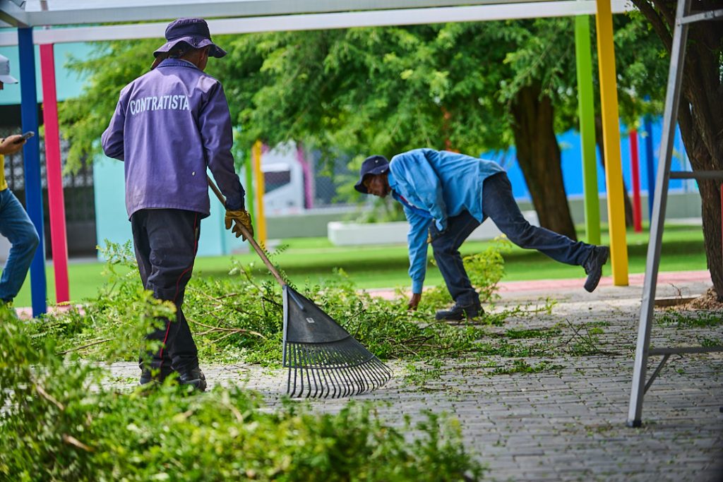 Dos trabajadores de mantenimiento con uniformes y sombreros recogen restos de poda y follaje en un parque de Palmira con estructuras de colores vivos al fondo