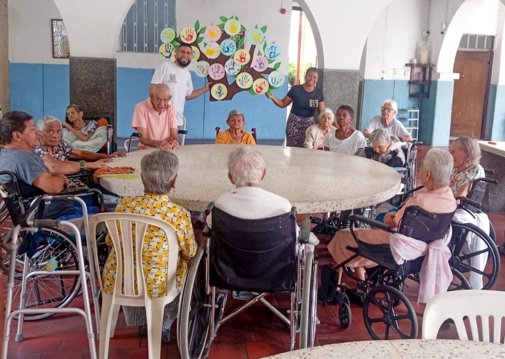 Un grupo de personas mayores y cuidadores se reúnen alrededor de una gran mesa circular en un centro de cuidado en Palmira. Al fondo, destaca un mural colorido de un árbol hecho con huellas de manos
