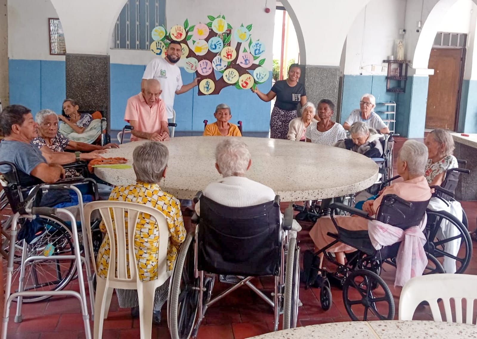 Un grupo de personas mayores y cuidadores se reúnen alrededor de una gran mesa circular en un centro de cuidado en Palmira. Al fondo, destaca un mural colorido de un árbol hecho con huellas de manos