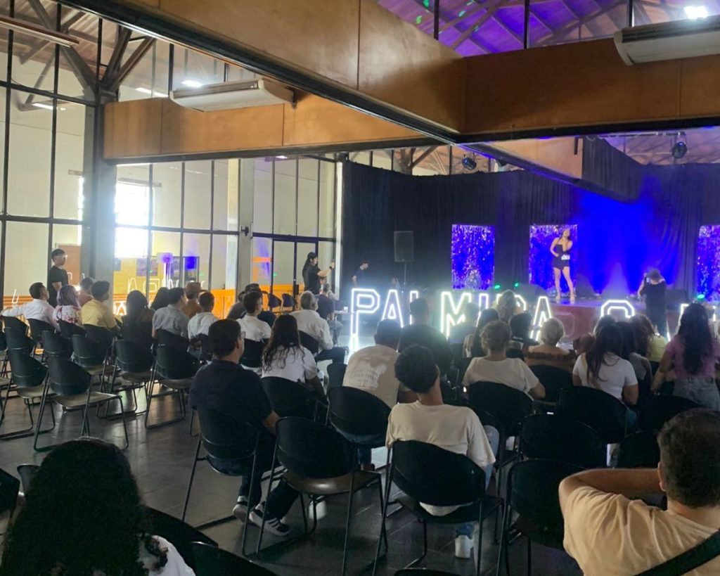 Una audiencia diversa y sentada observa una presentación musical en un escenario interior. Una gran señalización LED brillante deletrea "PALMIRA CANTA" frente al escenario.