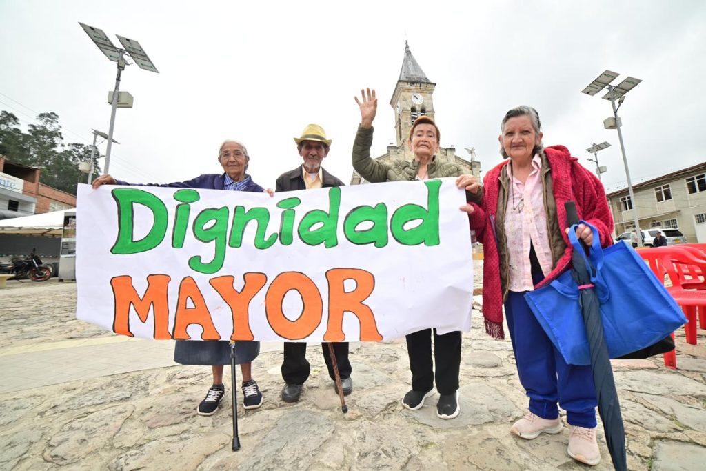 Cuatro personas mayores en una plaza pública sostienen una pancarta blanca con el mensaje "Dignidad MAYOR" en letras verdes y naranjas. Al fondo se observa una iglesia y postes de energía solar