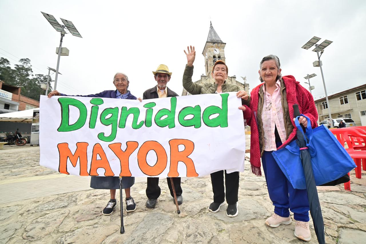 Cuatro personas mayores en una plaza pública sostienen una pancarta blanca con el mensaje "Dignidad MAYOR" en letras verdes y naranjas. Al fondo se observa una iglesia y postes de energía solar