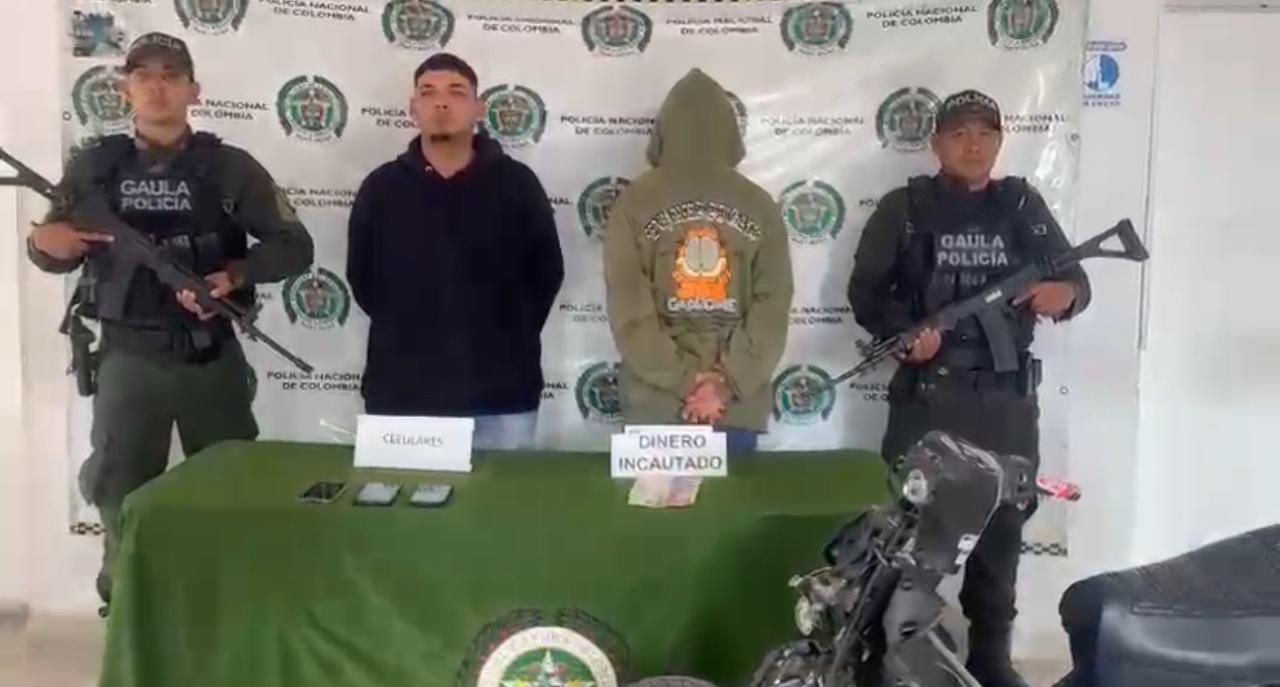 Dos integrantes de la unidad GAULA de la Policía Nacional custodian a dos personas bajo investigación. Al frente, una mesa con elementos incautados: teléfonos móviles, dinero en efectivo y una motocicleta. El fondo muestra logos institucionales de la Policía de Colombia.