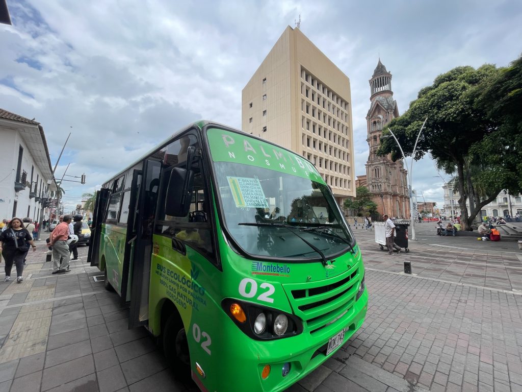 Bus ecológico de color verde vibrante estacionado en una plaza céntrica de Palmira perteneciente al sistema PALMIBUS. Al fondo, se observa arquitectura urbana, incluyendo una torre de la Catedral y personas transeúntes en una zona peatonal