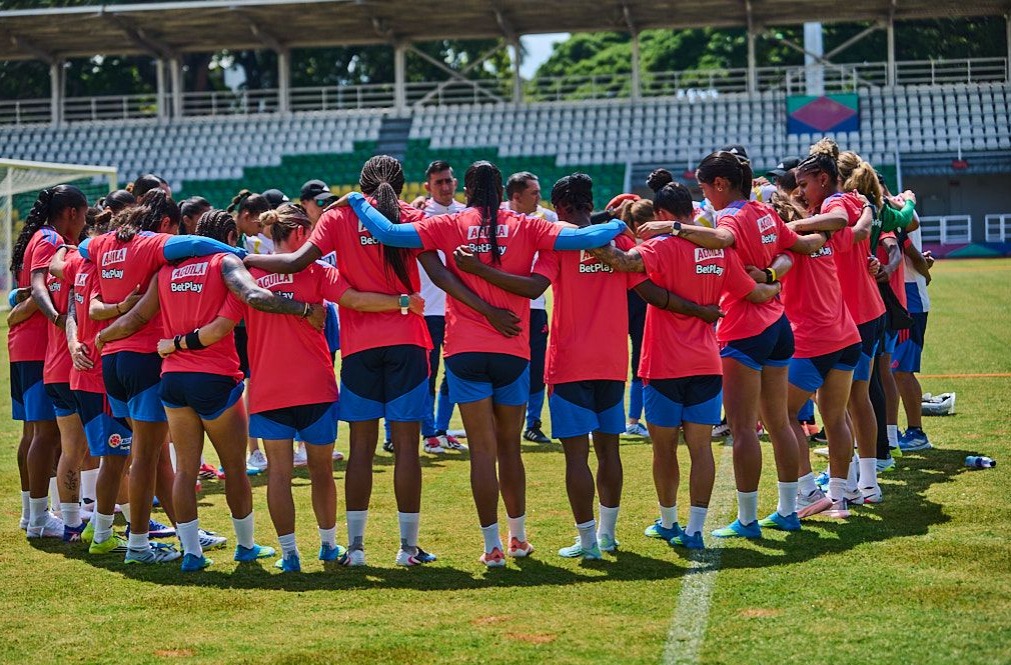 Jugadoras de fútbol de la selección colombiana en un círculo de unión durante un entrenamiento en el estadio. Visten uniformes rojos y azules, y se abrazan por los hombros en señal de compañerismo