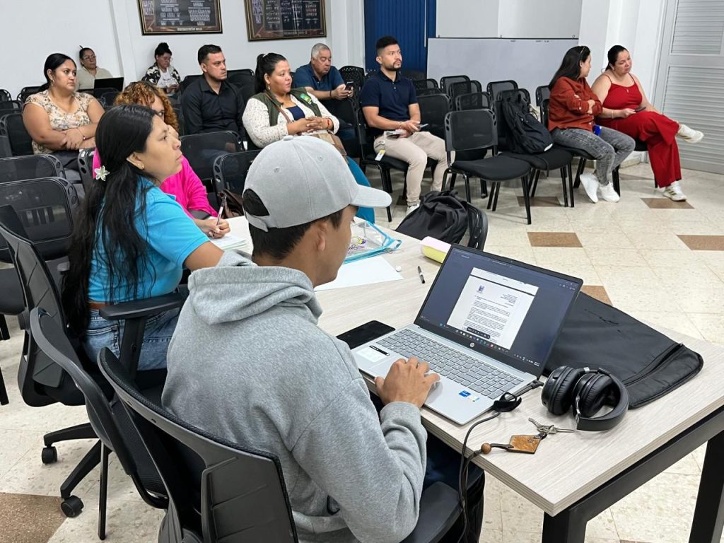 Personas en una jornada de trabajo y capacitación en un salón institucional. En primer plano, una persona con gorra gris utiliza un computador portátil, mientras al fondo un grupo diverso de asistentes escucha con atención