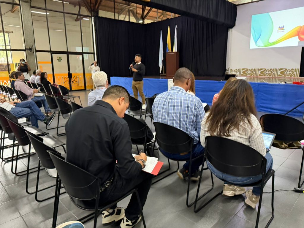 Una persona joven se dirige desde el centro de un auditorio a un grupo de asistentes sentados, quienes toman notas y escuchan atentamente. Al fondo, una tarima con banderas y una pantalla con el escudo de la Alcaldía de Palmira.