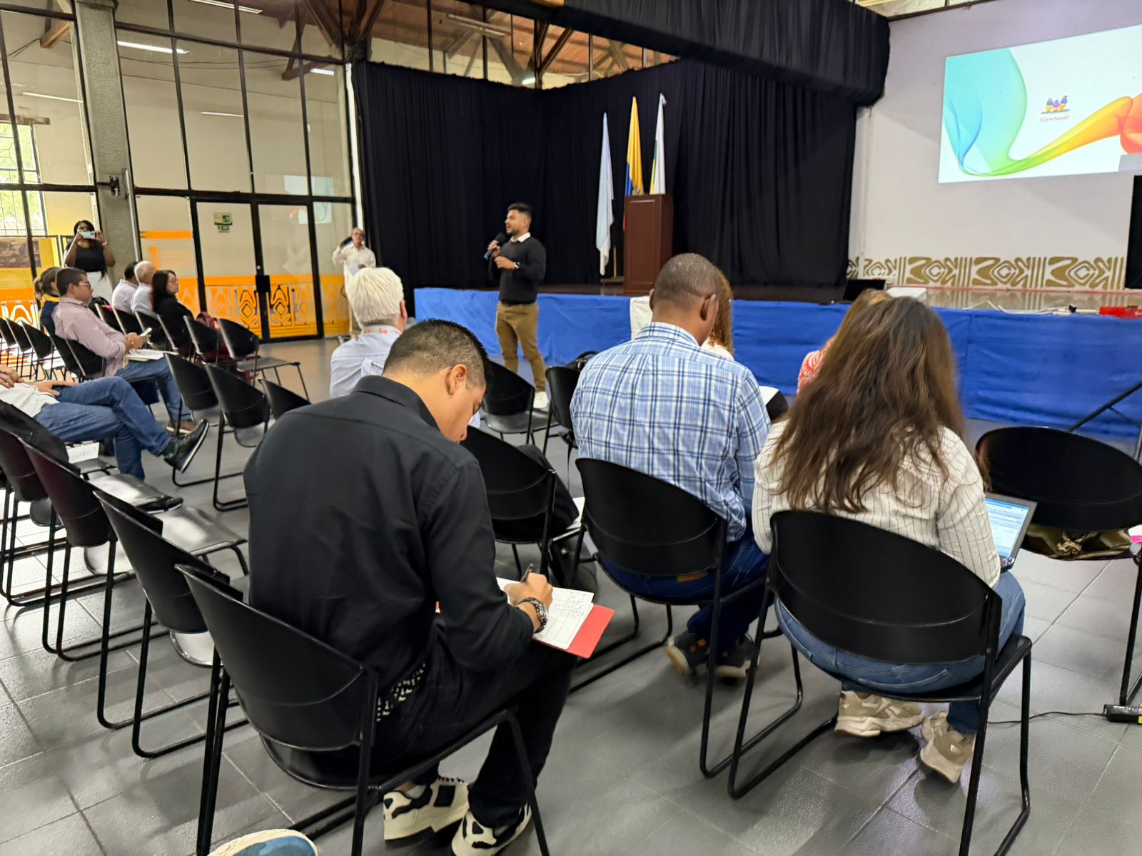 Una persona joven se dirige desde el centro de un auditorio a un grupo de asistentes sentados, quienes toman notas y escuchan atentamente. Al fondo, una tarima con banderas y una pantalla con el escudo de la Alcaldía de Palmira.