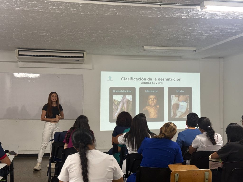Una mujer joven expone frente a un grupo de estudiantes en un salón de clases. En la pantalla proyectada se lee "Clasificación de la desnutrición aguda severa" con ejemplos de Kwashiorkor, Marasmo y Mixta