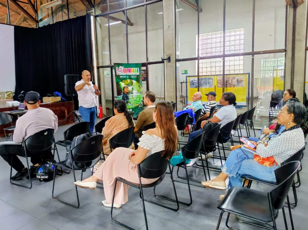 Persona ponente dirigiéndose a un grupo de asistentes sentados en un salón. Al fondo se observa un cartel promocional de 'Vive el Valle' con imágenes de naturaleza