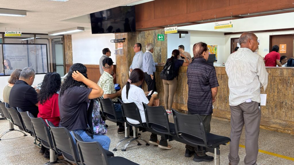 Ciudadanía palmirana de diversas edades espera su turno de atención en la Oficina del Contribuyente. Varias personas se encuentran sentadas en hileras de sillas oscuras, mientras otros ciudadanos y ciudadanas aguardan de pie frente a las ventanillas de trámites y pagos. El espacio es iluminado y cuenta con señalética clara para las casillas de servicio