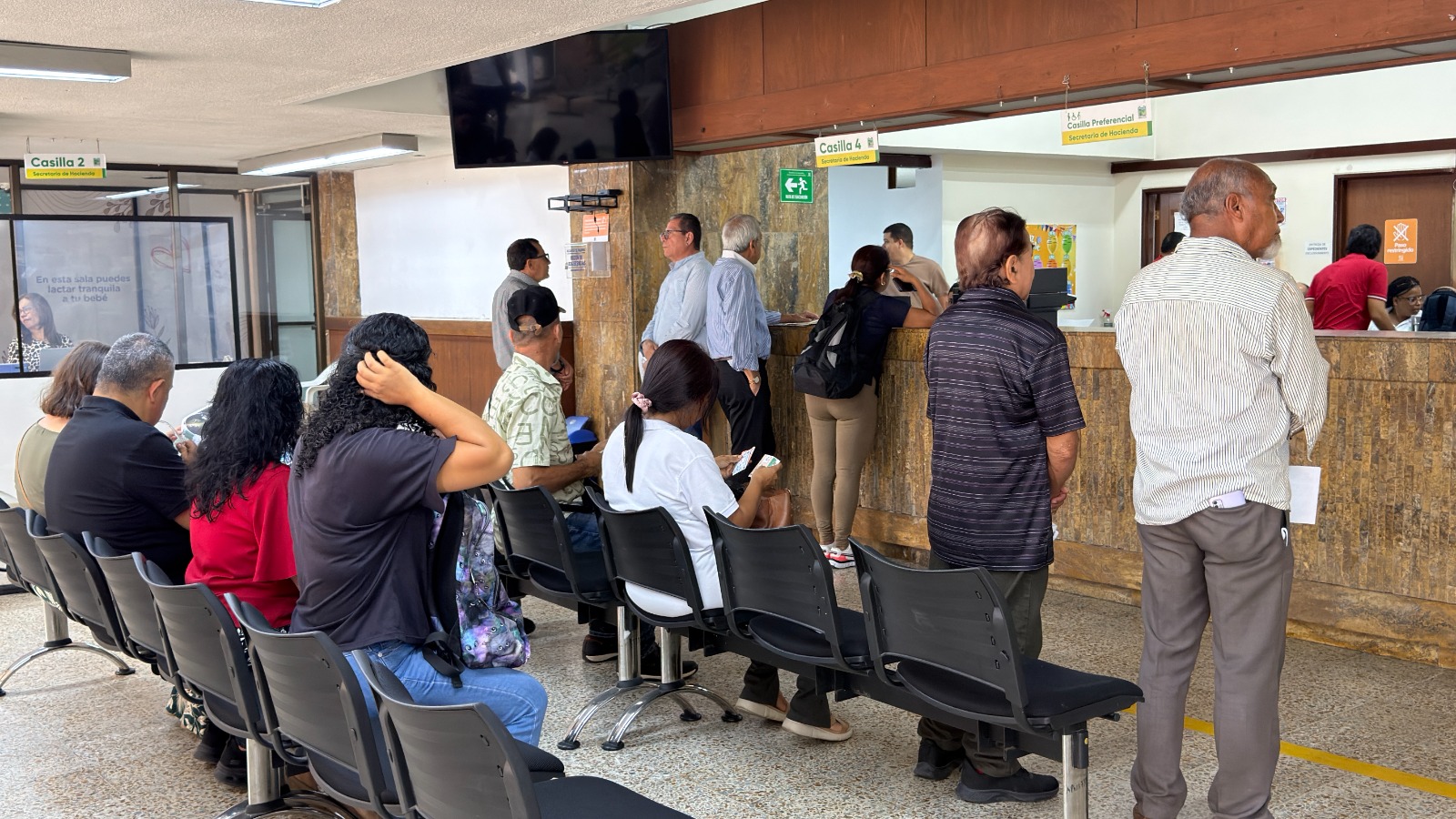 Ciudadanía palmirana de diversas edades espera su turno de atención en la Oficina del Contribuyente. Varias personas se encuentran sentadas en hileras de sillas oscuras, mientras otros ciudadanos y ciudadanas aguardan de pie frente a las ventanillas de trámites y pagos. El espacio es iluminado y cuenta con señalética clara para las casillas de servicio