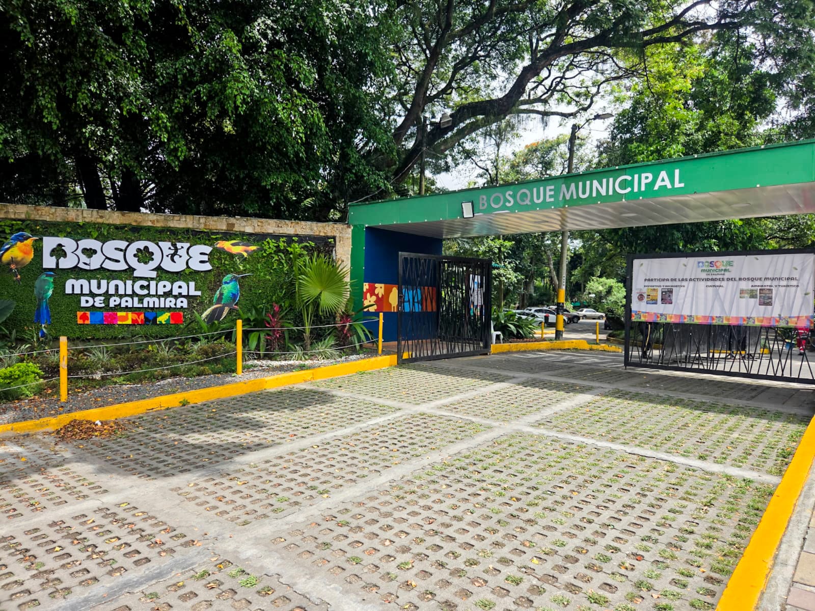 Entrada principal del Bosque Municipal de Palmira. Se ve un letrero colorido con aves, una gran valla verde que dice 'Bosque Municipal' y un área de estacionamiento con bloques de césped