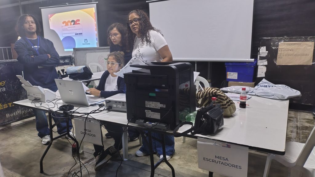 Equipo técnico trabajando en una mesa con equipos de cómputo e impresión. Se observan letreros que indican 'Mesa Escrutadores' y 'Mesa Digitador' en un entorno de oficina o centro de votación