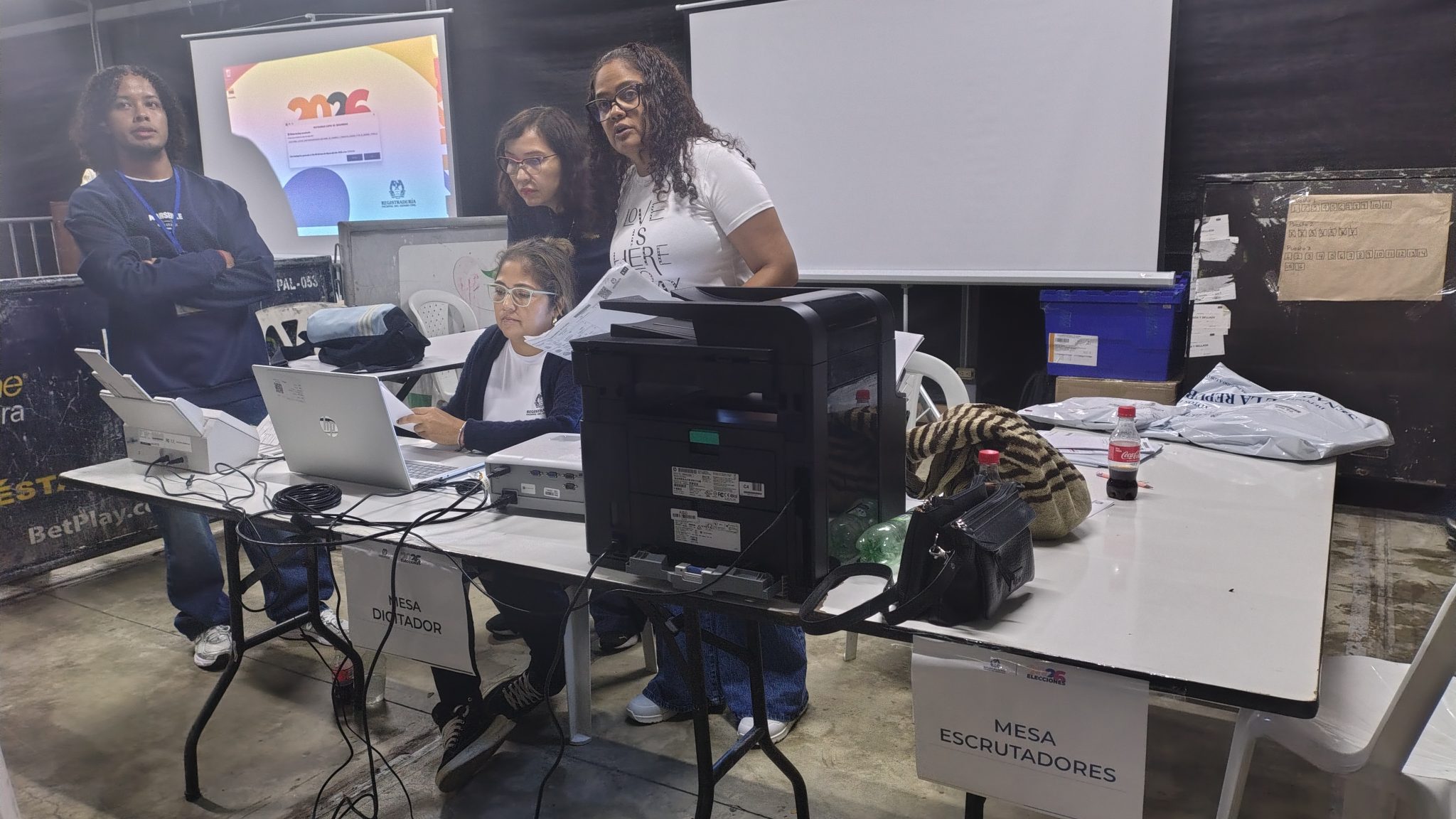 Equipo técnico trabajando en una mesa con equipos de cómputo e impresión. Se observan letreros que indican 'Mesa Escrutadores' y 'Mesa Digitador' en un entorno de oficina o centro de votación