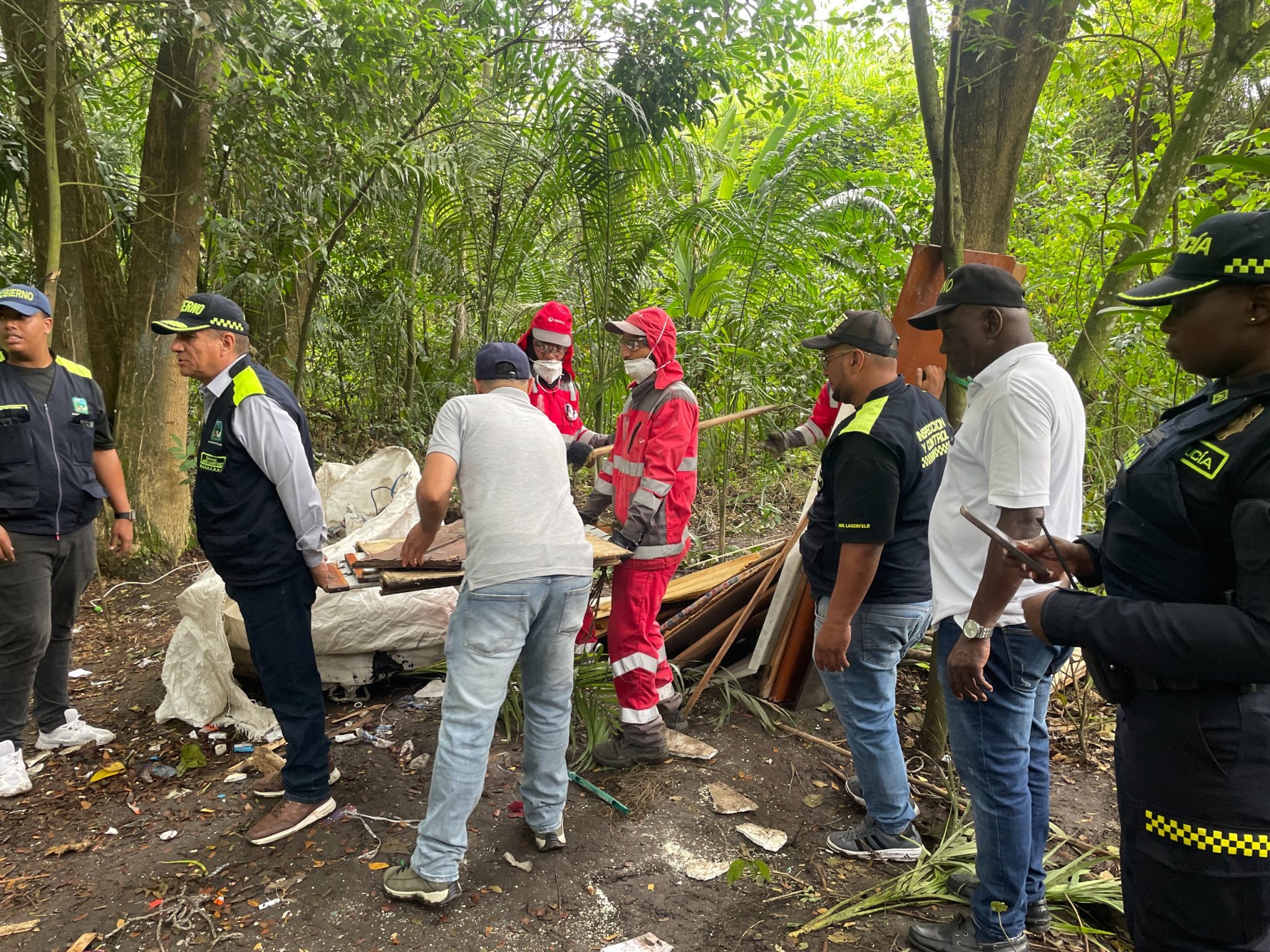 Grupo de personas con uniformes de organismos de socorro, policía y vestimenta civil trabajando en la remoción de materiales en un entorno natural boscoso