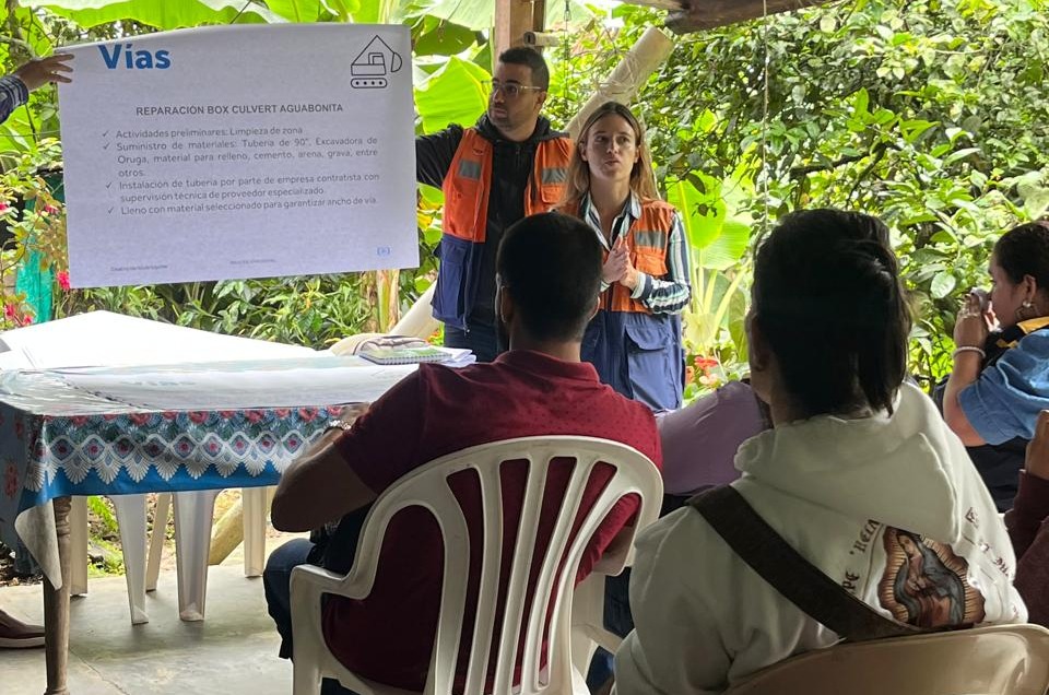 Personal de la administración municipal presenta un cartel informativo sobre la reparación de la vía Aguabonita ante un grupo de personas de la comunidad en un espacio abierto y arbolado