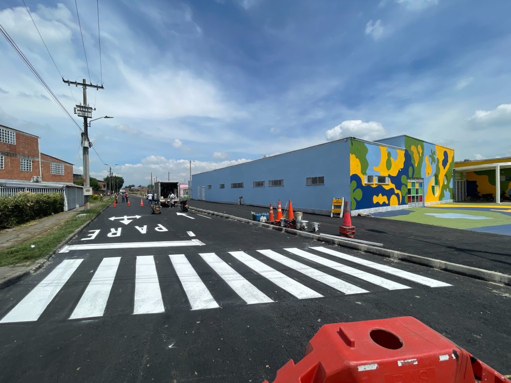 Fotografía de una calle recién pavimentada frente a un edificio colorido en Palmira. Se observa un paso peatonal pintado, la señal de 'PARE' en el asfalto y personal trabajando en el mantenimiento vial.