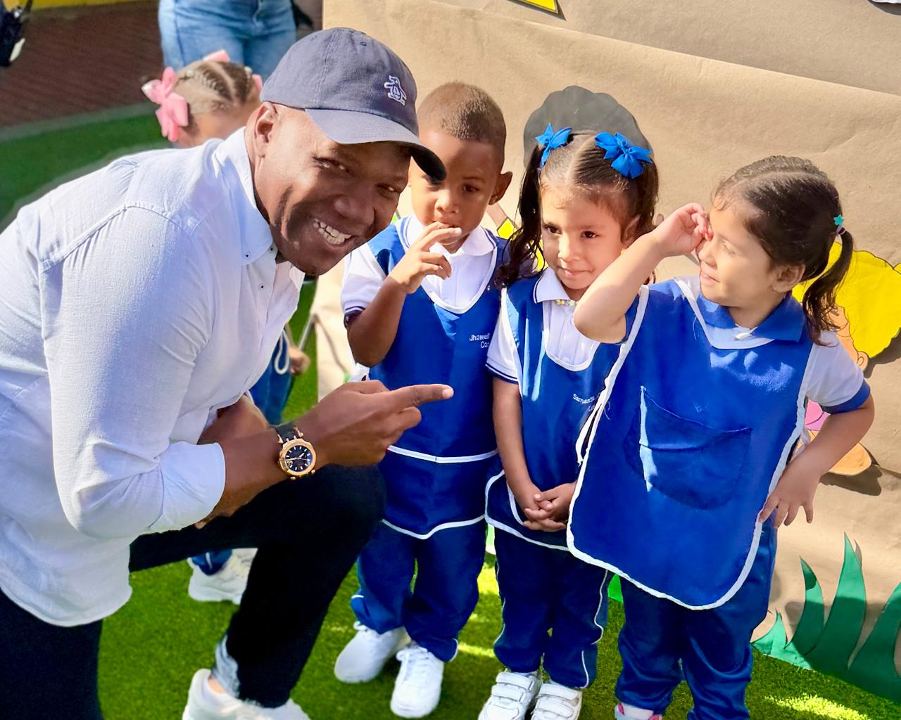 El alcalde de Palmira, Víctor Ramos, sonríe y señala a tres niños y niñas de primera infancia que visten uniformes escolares azules. El grupo se encuentra en un espacio educativo al aire libre decorado con motivos infantiles
