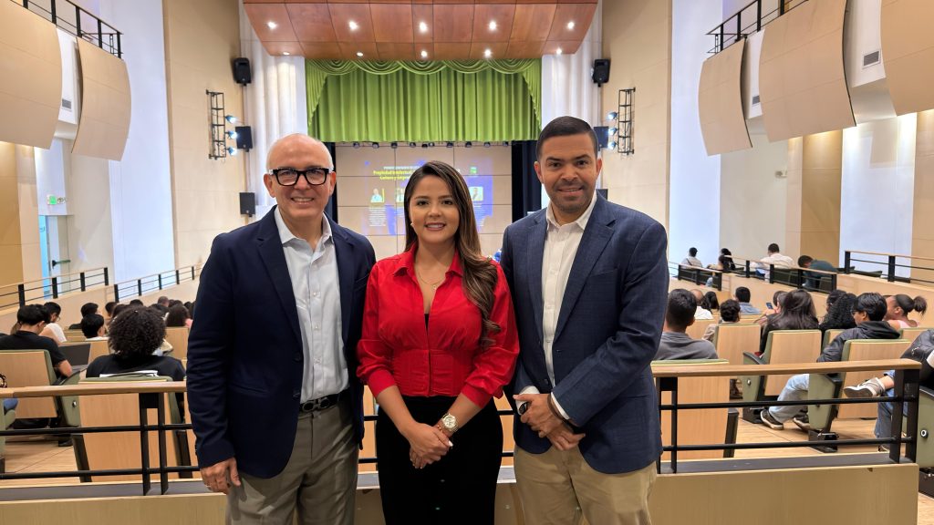 Tres personas profesionales (dos hombres y una mujer) posan sonriendo en la parte delantera de un auditorio moderno y bien iluminado. Detrás de ellos, se ven filas de asientos con algunas personas de la audiencia. En el escenario, hay una pantalla de proyección con texto e imágenes y una cortina verde.
