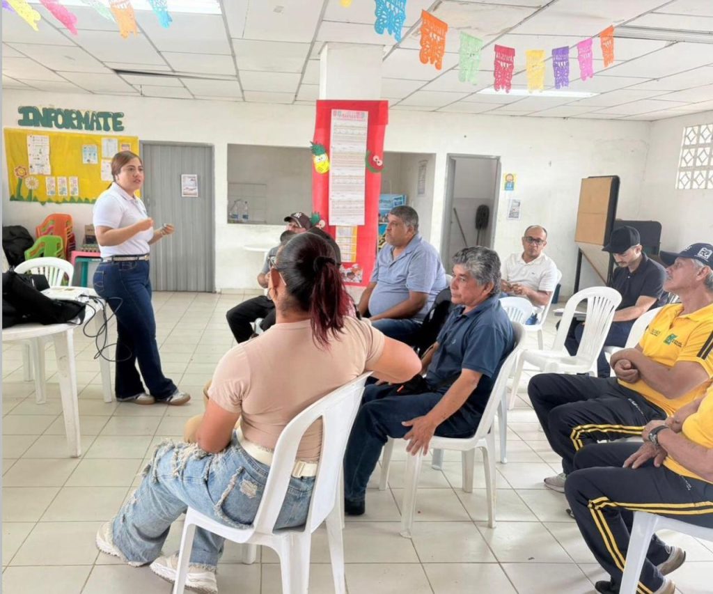 Una mujer joven con polo blanco y jeans expone frente a un grupo de personas sentadas en un salón decorado con papel picado. El ambiente es de una charla informativa o comunitaria