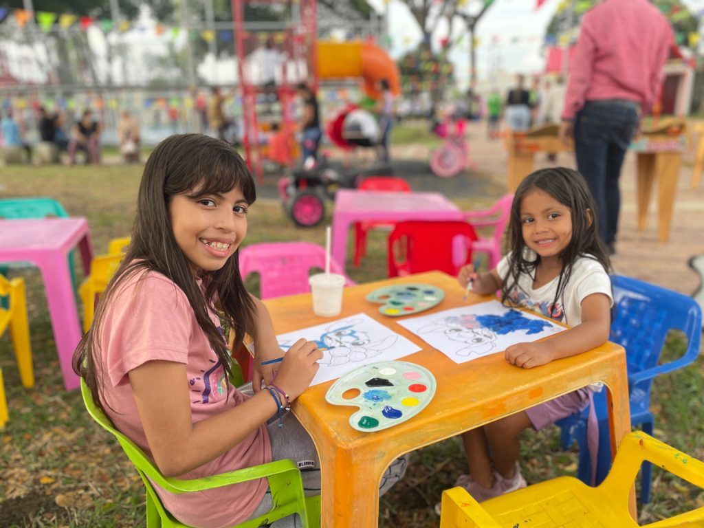 Dos personas menores de edad están sentadas en una pequeña mesa naranja en un parque, cada una coloreando dibujos de personajes de Stitch con pinceles y paletas de pintura. Miran a la cámara y sonríen. El fondo está desenfocado, mostrando un área de juego al aire libre con más mesas de colores y personas adultas borrosas