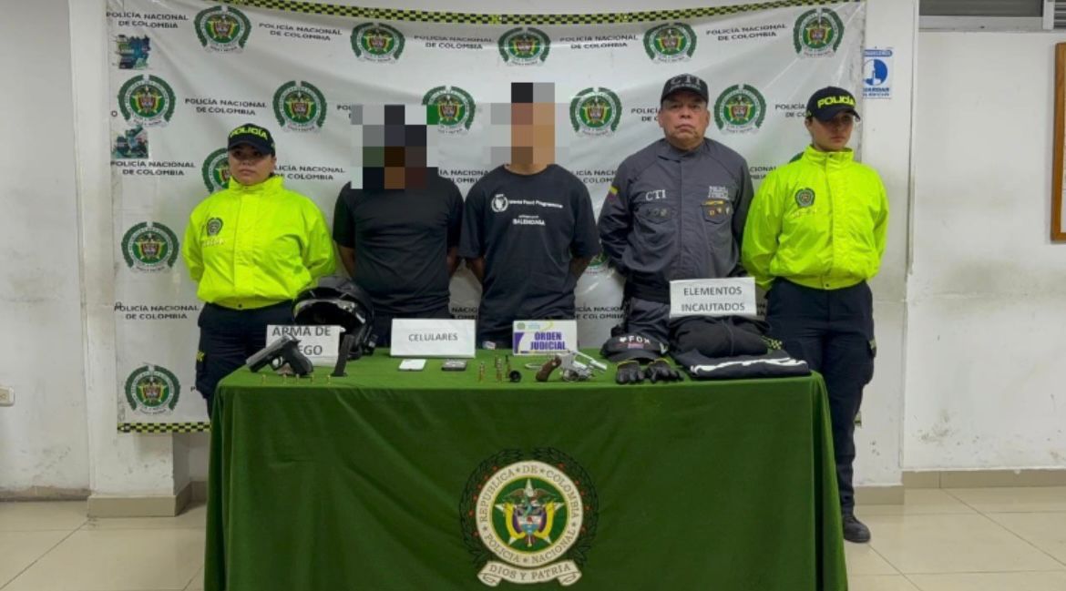 Integrantes de la Policía Nacional y el CTI junto a dos personas detenidas. Delante de ellos hay una mesa con elementos incautados como armas de fuego y celulares, bajo un fondo con logos oficiales