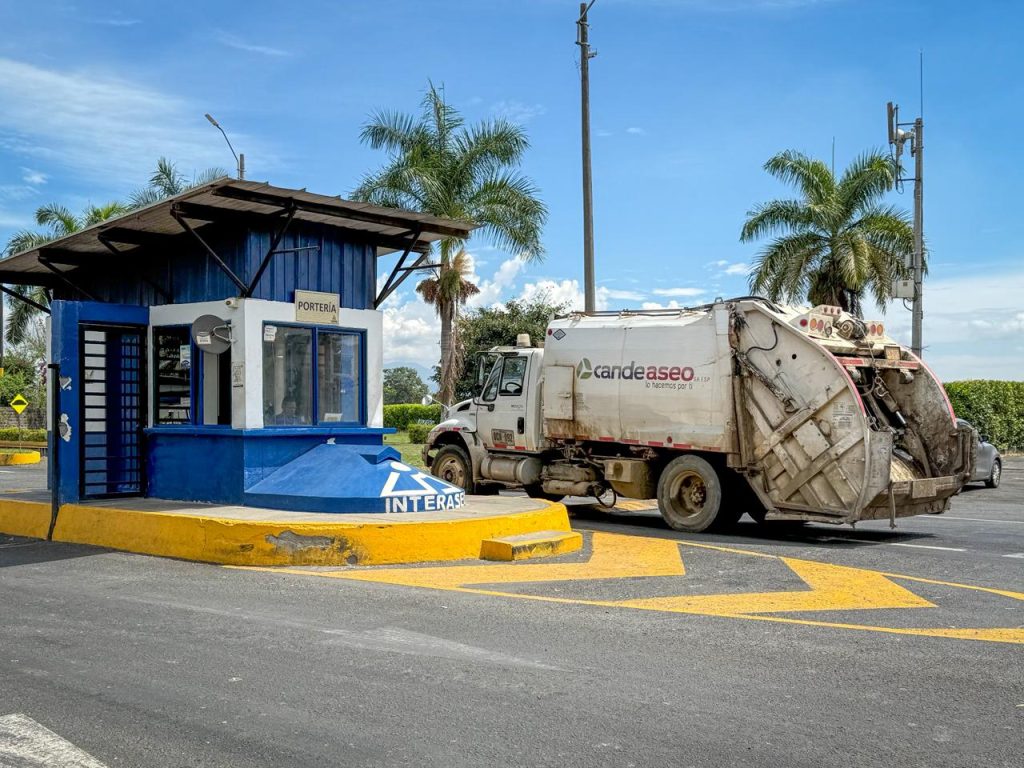Camión de recolección pasando junto a una caseta de vigilancia azul y blanca con el logo de Interaseo en Palmira, bajo un cielo despejado