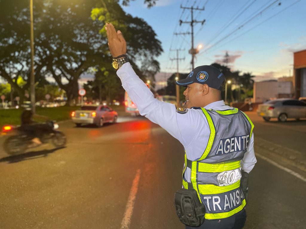 Fotografía de perfil de un agente de tránsito en Palmira durante la jornada nocturna. Viste uniforme oficial con chaleco reflectante y realiza señales manuales para regular el flujo vehicular en una vía principal de la ciudad.