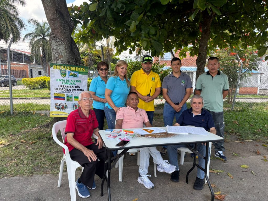 otografía de un grupo de líderes y lideresas de la Junta de Acción Comunal de la Urbanización Ciudadela Palmira. Se encuentran reunidos bajo la sombra de un árbol en una jornada de participación ciudadana; algunos están sentados frente a una mesa con planillas de registro y otros permanecen de pie junto a un pendón informativo de la JAC.